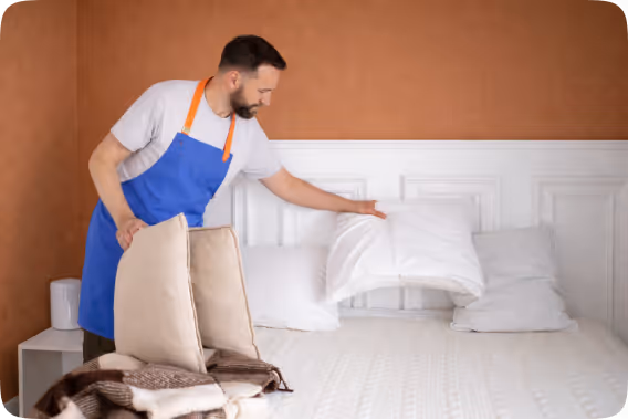 Man in blue apron arranging pillows on a bed with a white headboard and beige wall.