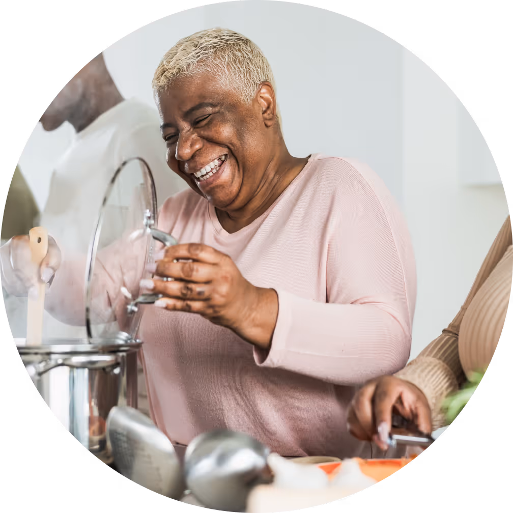 A smiling woman in a light pink shirt holds the lid of a pot while cooking in a kitchen, surrounded by others who are preparing food.