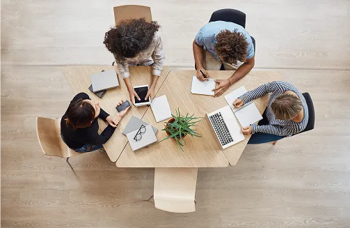 Quatre personnes assises autour d'une table triangulaire en bois, utilisant un ordinateur portable, une tablette, des carnets et des smartphones.