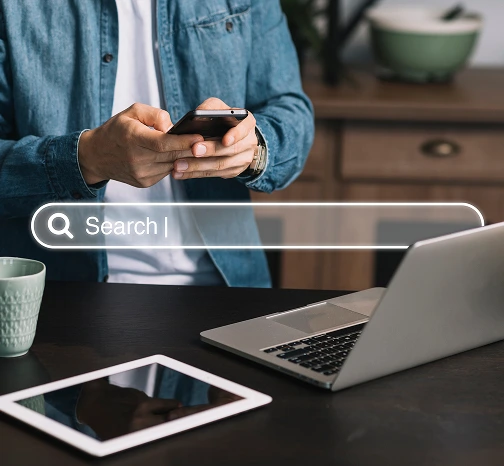 Personne utilisant un smartphone avec un ordinateur portable et une tablette sur une table, illustration d'une barre de recherche virtuelle.