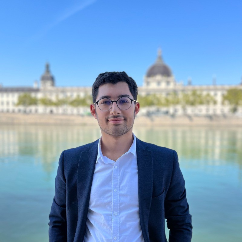 Photo de Luca Giustino, le fondateur de l'agence 998. Au premier plan, Luca apparaît en costume et chemise. En arrière-plan se distinguent un fleuve et un bâtiment historique.