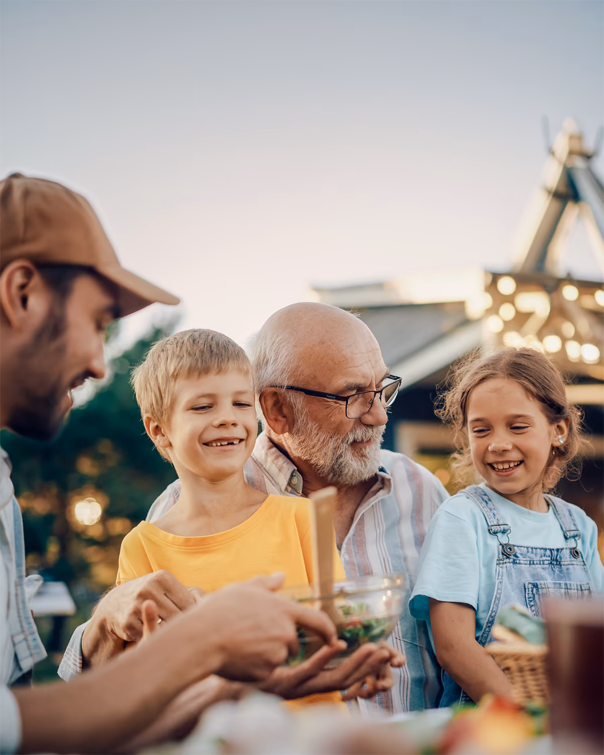A multigenerational family — a young father, two children, and a smiling older man with a gray beard — gathered around a table outdoors at dusk