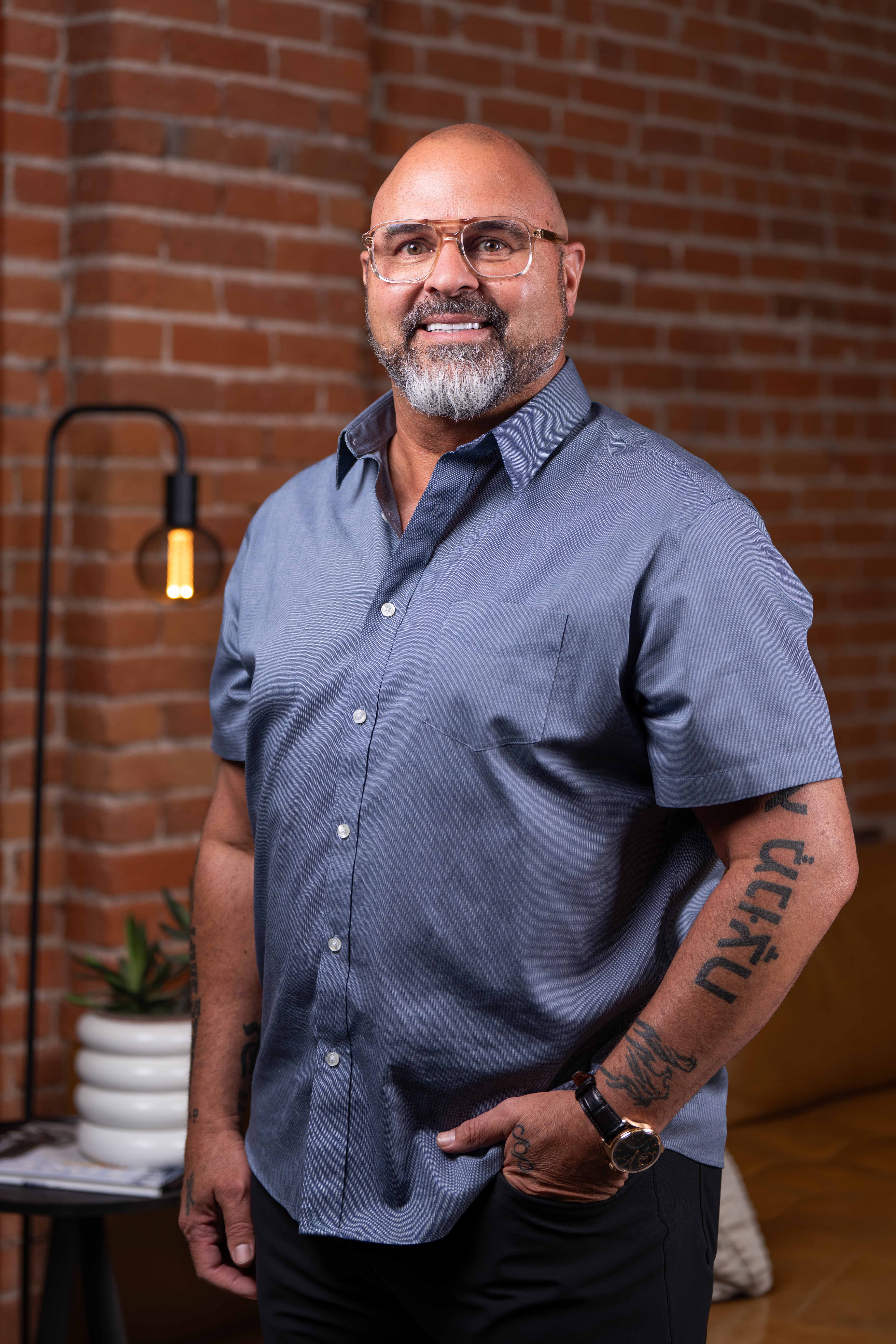 Dr. Kirk Elliott smiling in a blue button-up shirt, standing in front of an exposed brick wall