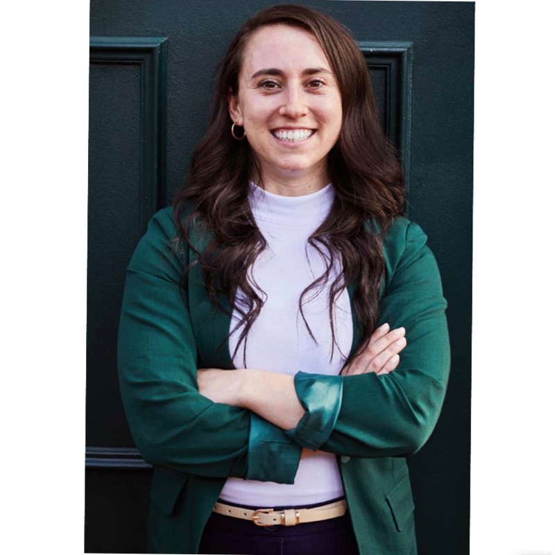 Smiling woman with long brown hair, wearing a green jacket and white turtleneck, standing with arms crossed against a dark door.