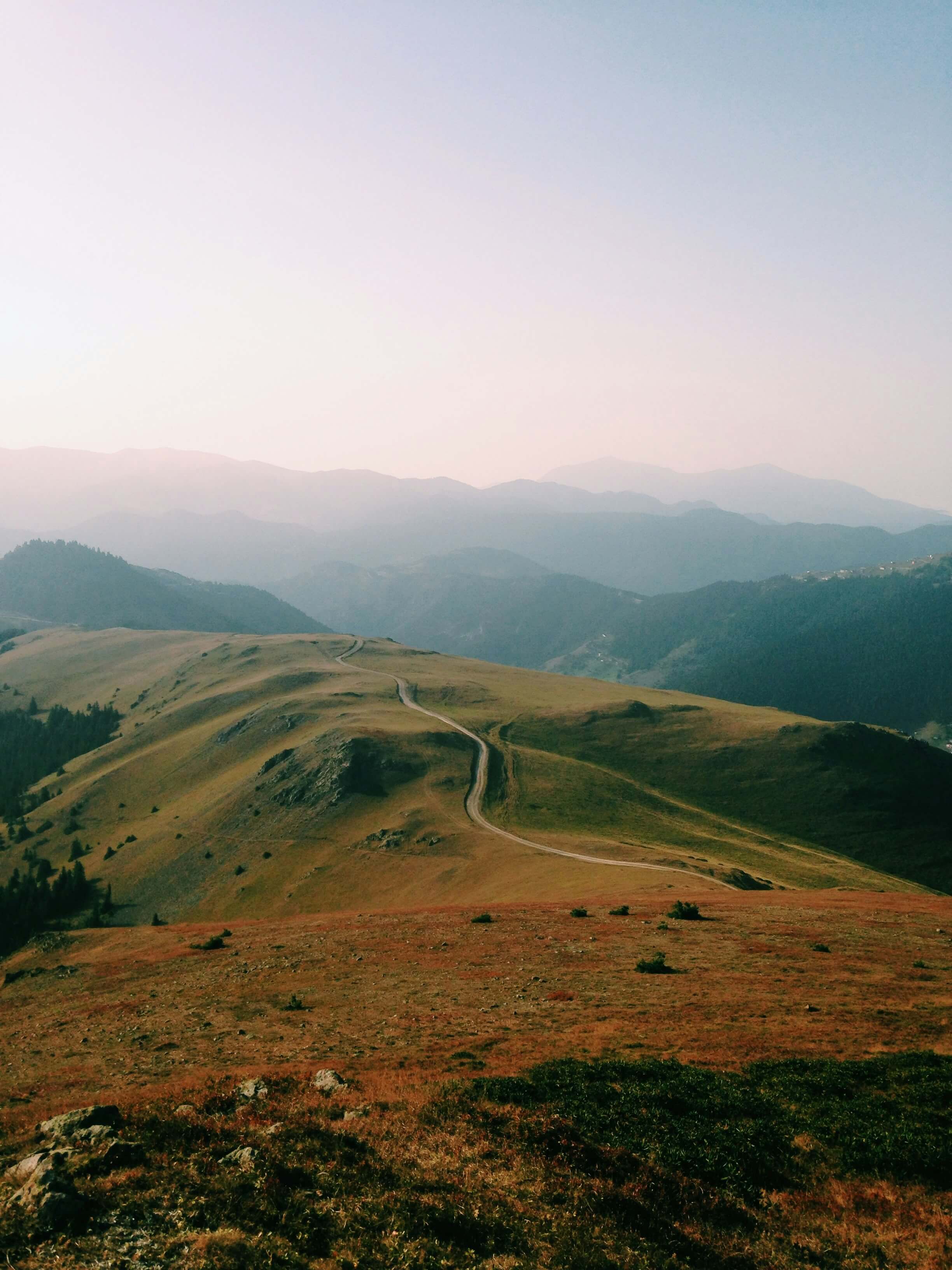 Rolling hills with a winding dirt path under a hazy sky and layers of distant mountains.