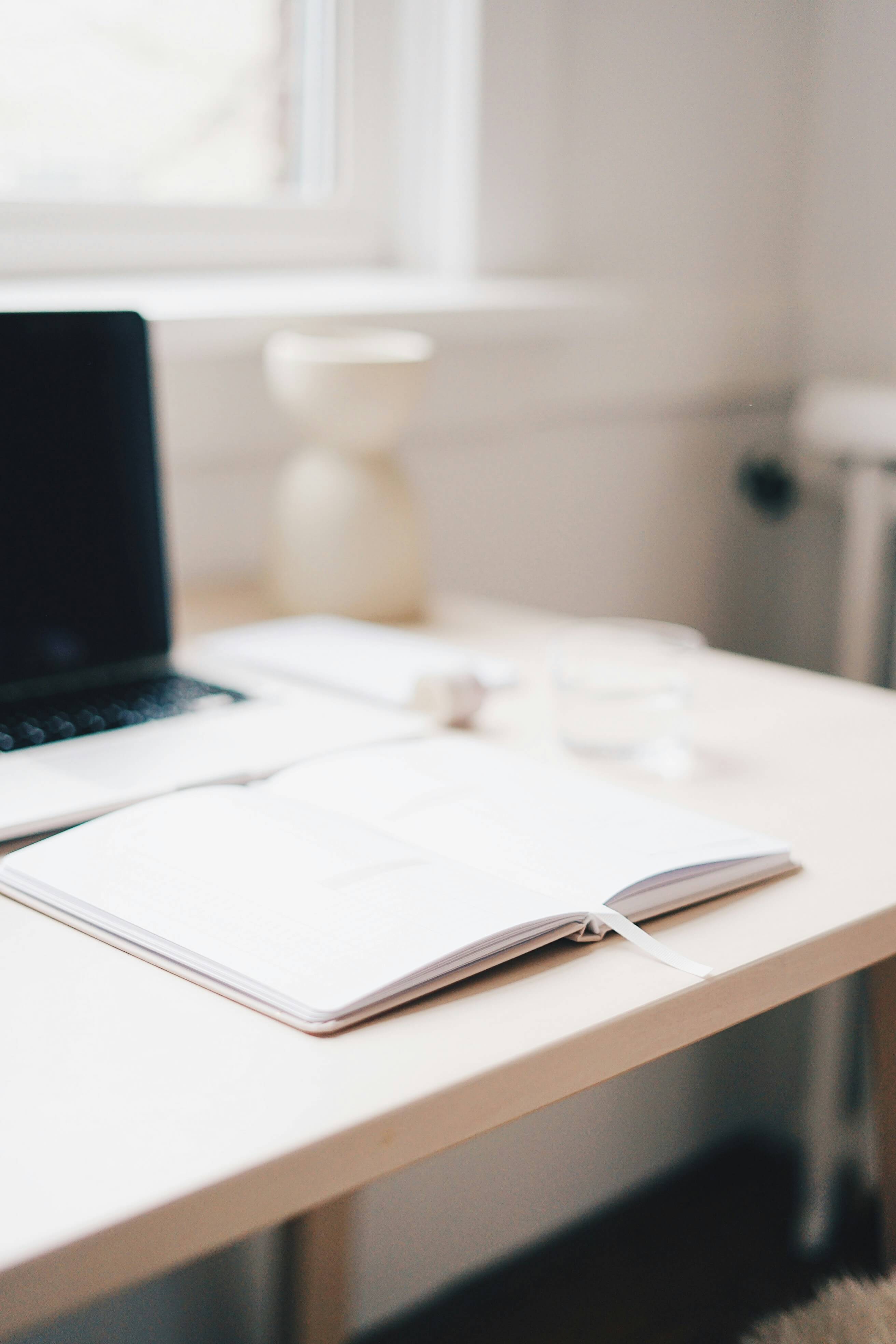Open notebook with a ribbon bookmark on a light wooden desk with a laptop and a glass of water in soft focus background.