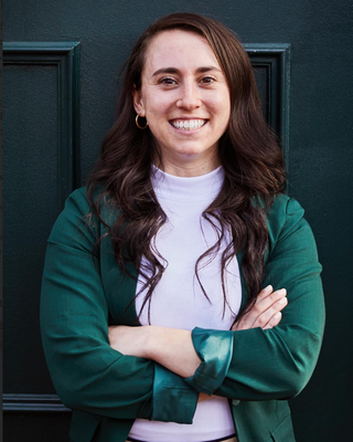 Smiling woman with long brown hair, wearing a green blazer and white shirt, stands with arms crossed in front of a dark door.