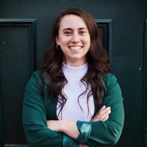 Smiling woman with long dark hair wearing a green jacket and white top, standing with arms crossed against a dark door background.