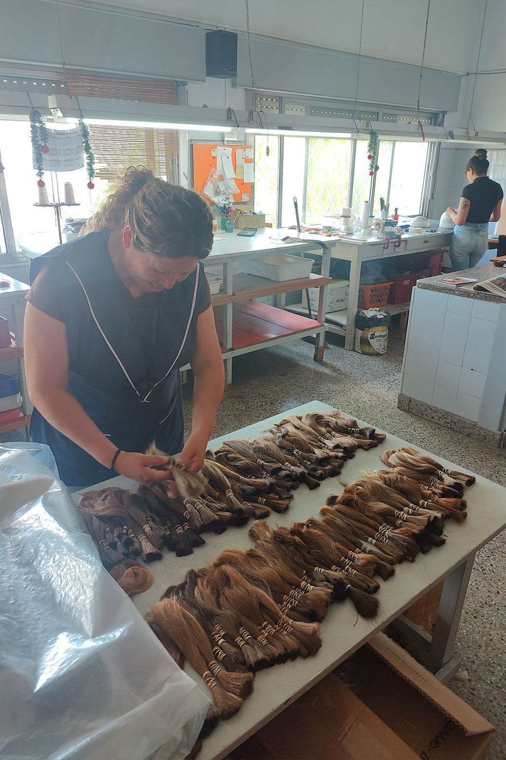 Woman in a workshop arranging bundles of brown hair extensions on a table, with another person working in the background.