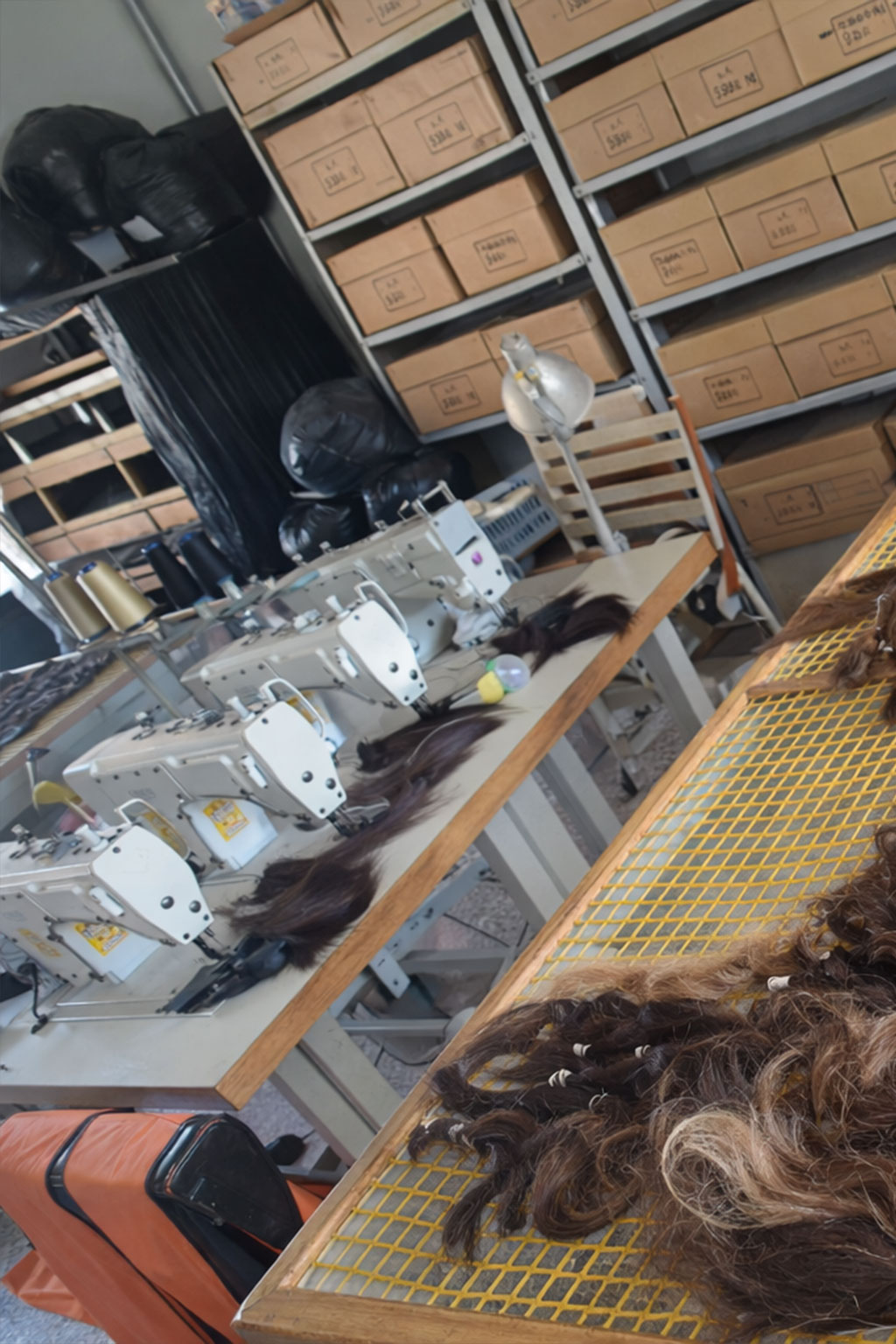 Workroom with multiple sewing machines, synthetic hair strands on tables, shelves filled with labeled cardboard boxes, and large black bags in the background.