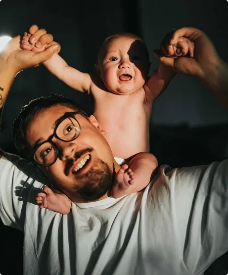 Smiling man wearing glasses holding a happy baby on his shoulders, with the baby raising arms and holding the man's hands.