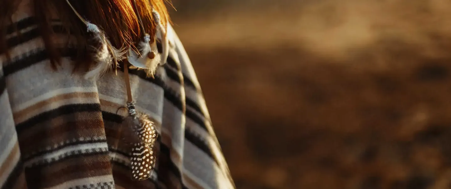 Close-up of a person wearing a striped poncho with a hanging feather accessory against a blurred brown background.