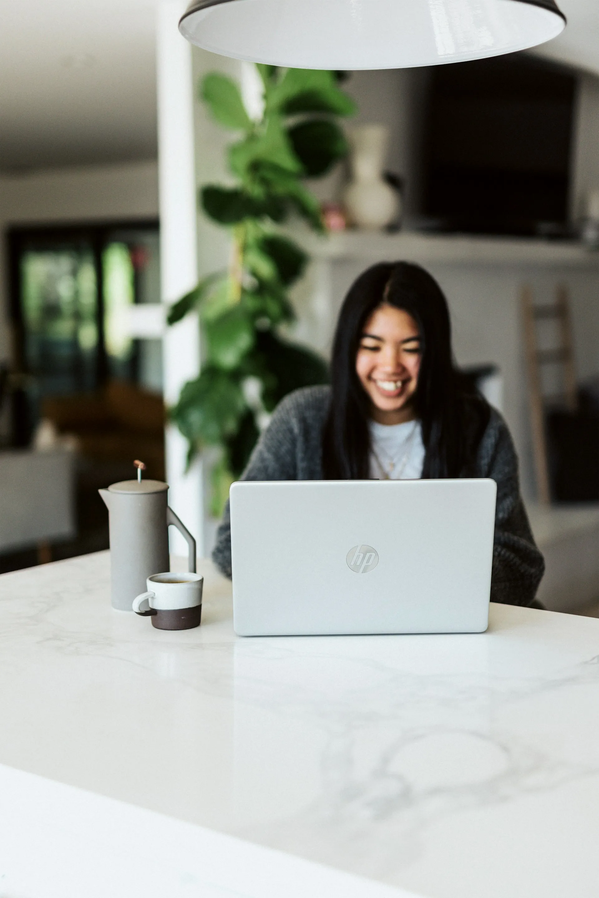 Smiling woman with long black hair working on a silver HP laptop at a white marble table with a coffee pot and cup.