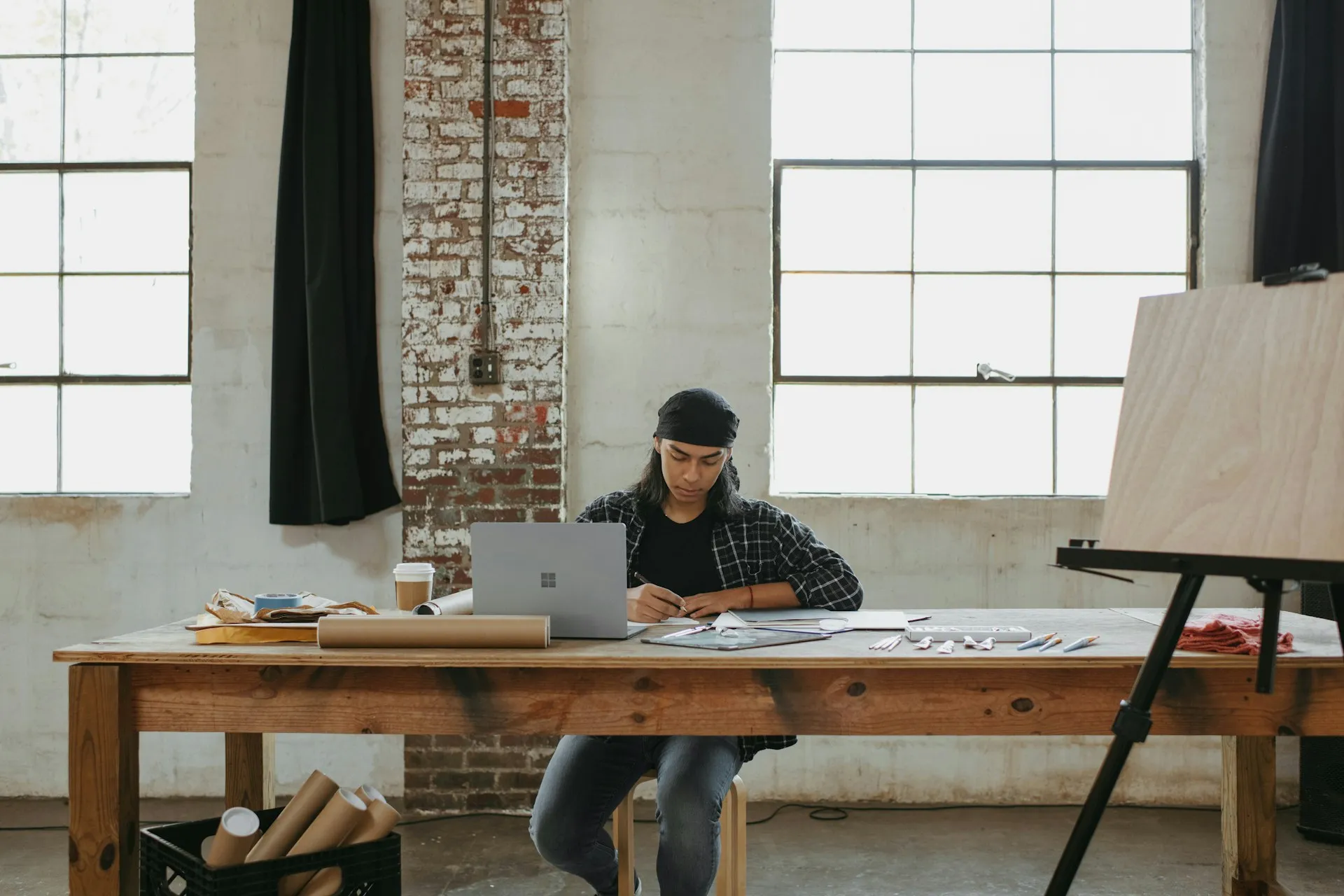 Person wearing a black bandana and plaid shirt working at a wooden table with a laptop and art supplies in a bright studio with large windows.