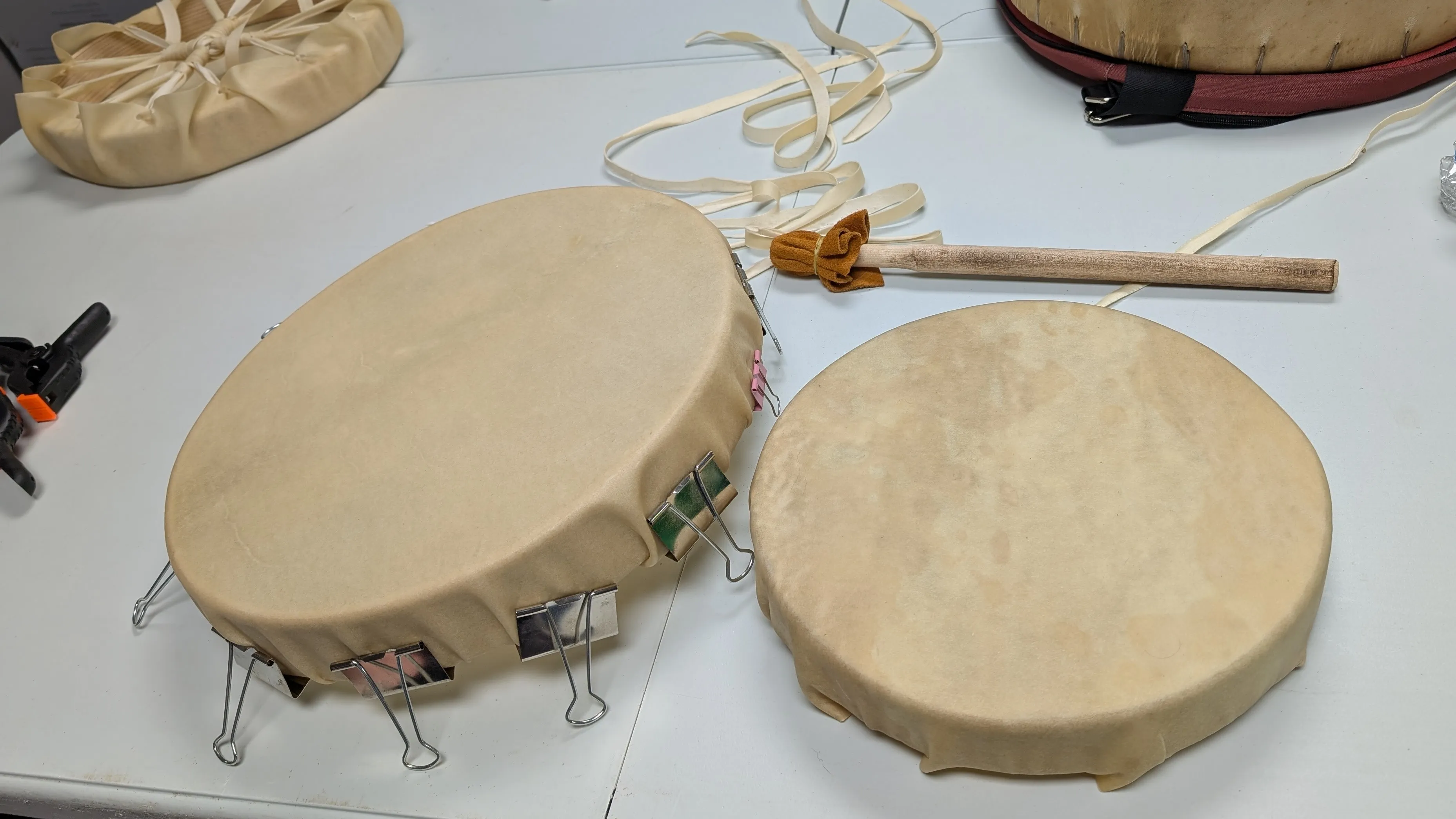 Two traditional hand drums with beige drumheads on a white surface, one secured with binder clips and the other with tied ribbons, along with a wooden drumstick wrapped with fabric.