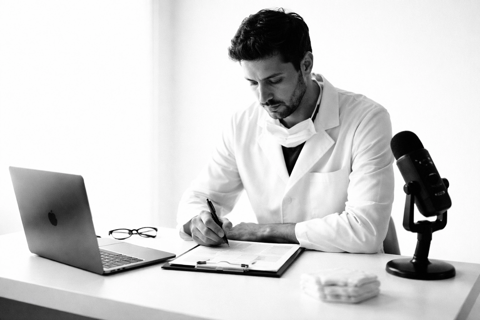 High-contrast black and white documentary photo of a doctor speaking into a podcast microphone in a minimal studio.