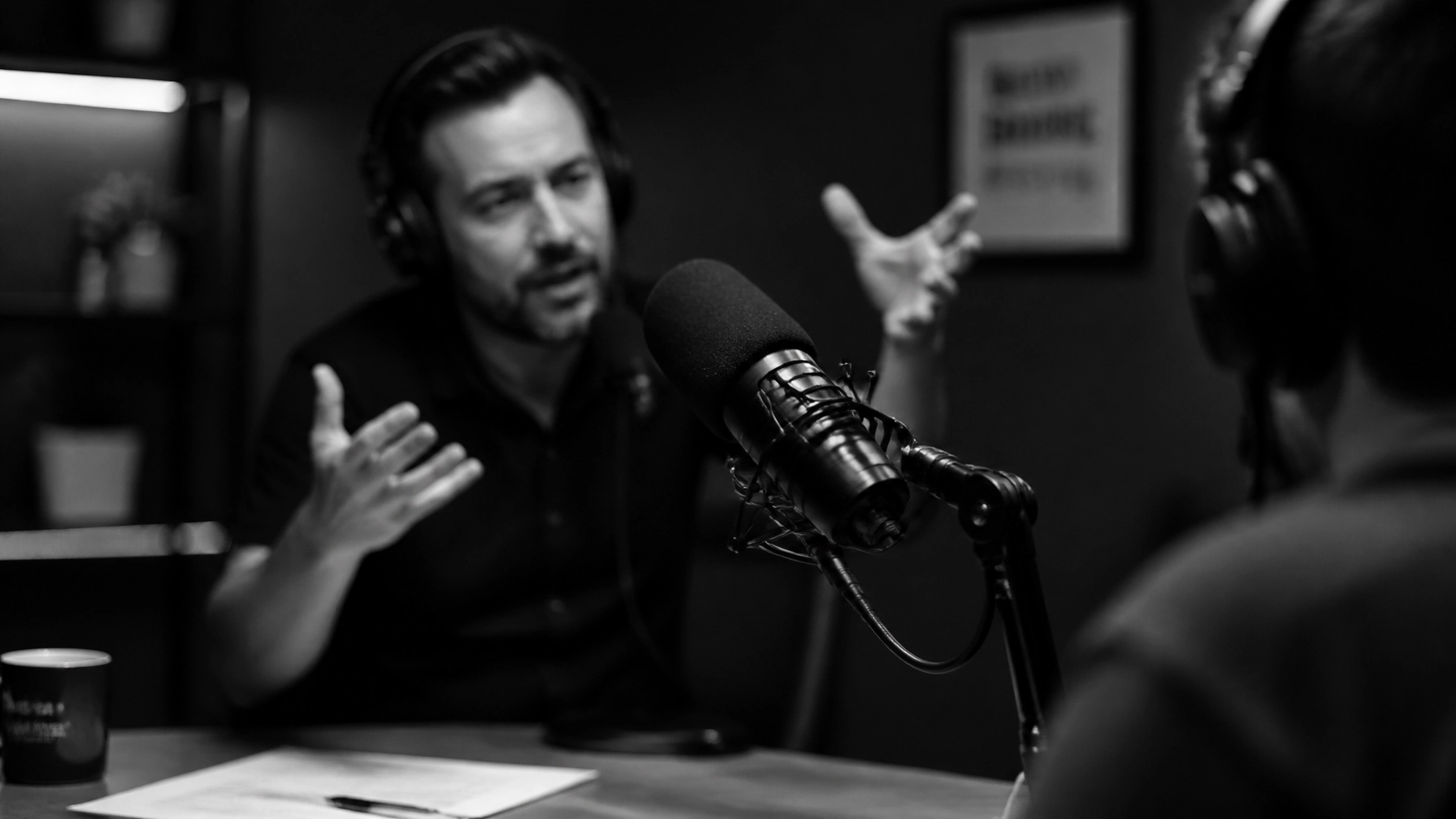 A high-contrast black and white candid photo of a professional podcast recording session. A microphone in focus with a blurry background of a person speaking passionately. Minimalist aesthetic.