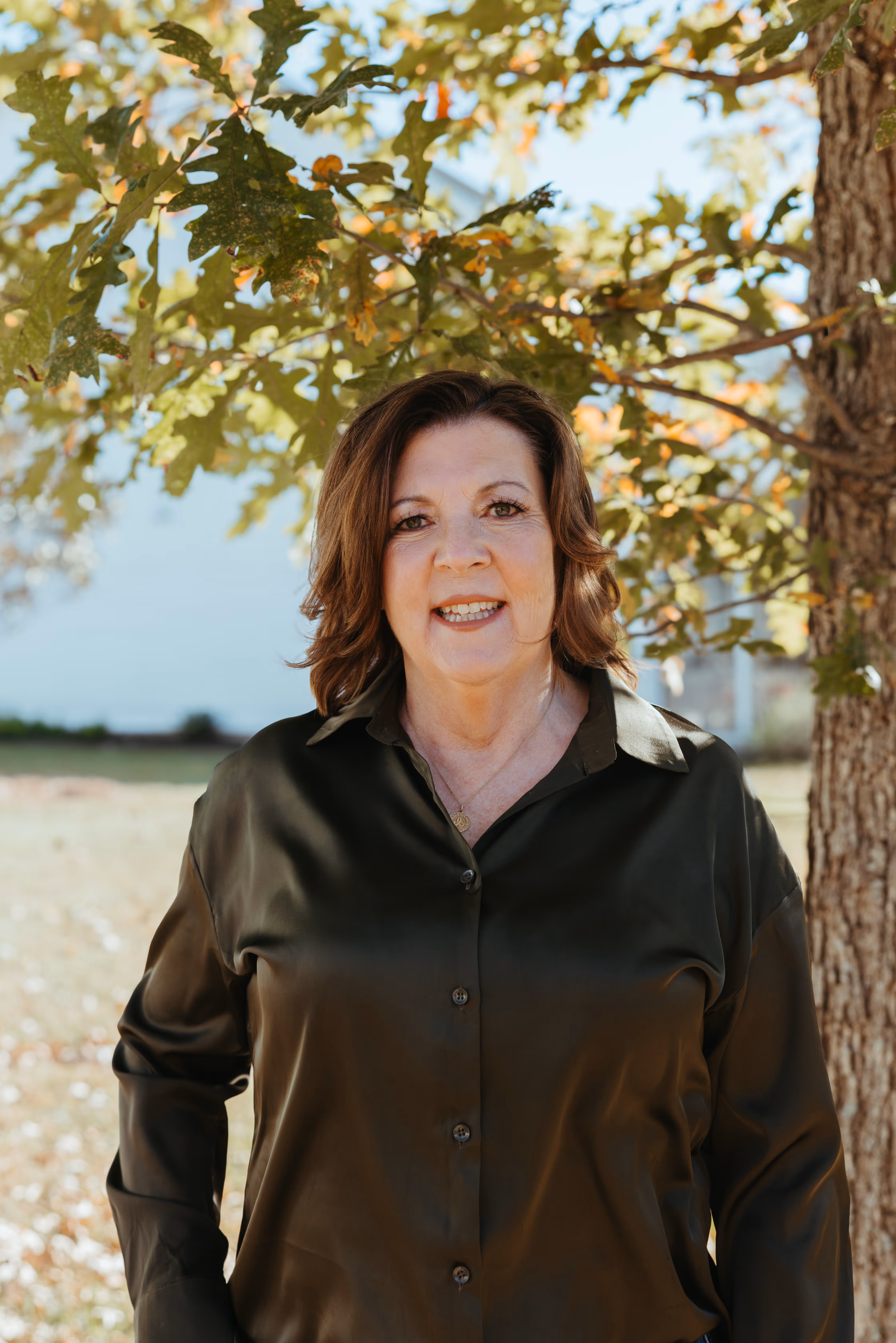 Mary Harkins, Board-Certified Educational Planner and Expert College Admissions Consultant at Gemsight stands outside, wearing a dark brown buttoned blouse, under tree branches with green leaves.
