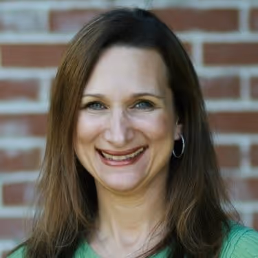 Jessica Sharkey, Expert College Essay Editor and Admissions Consultant at Gemsight is wearing a green top and hoop earrings against a brick wall background.