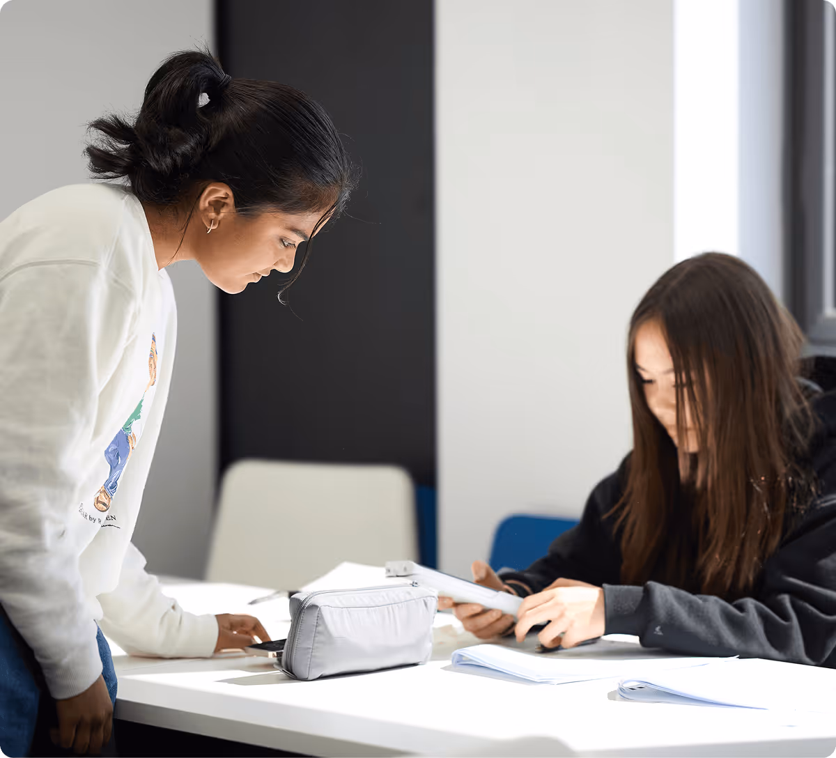 Two young women studying together at a white table with books and a pencil case in a bright room.