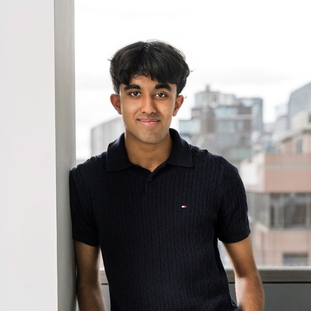 Young man with dark hair wearing a black polo shirt, leaning against a white wall with a blurred cityscape in the background.
