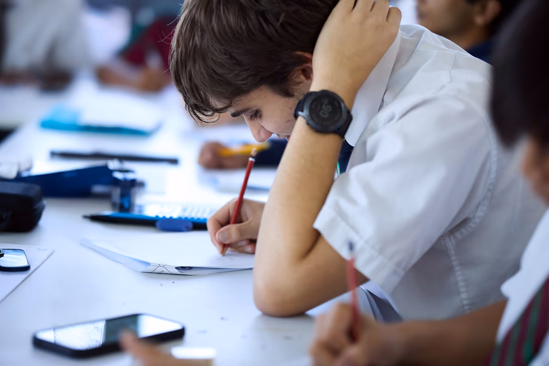 Student in a white shirt and watch writing with a pencil at a desk in a classroom.