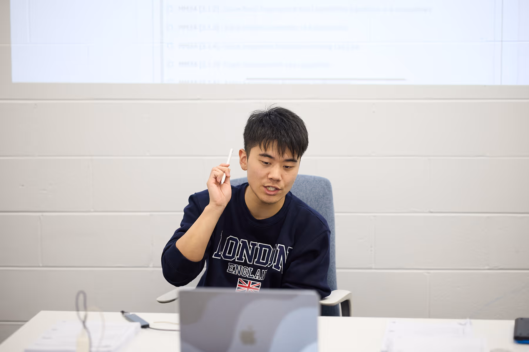Young man in a London sweatshirt holding a pen and speaking in front of a laptop.