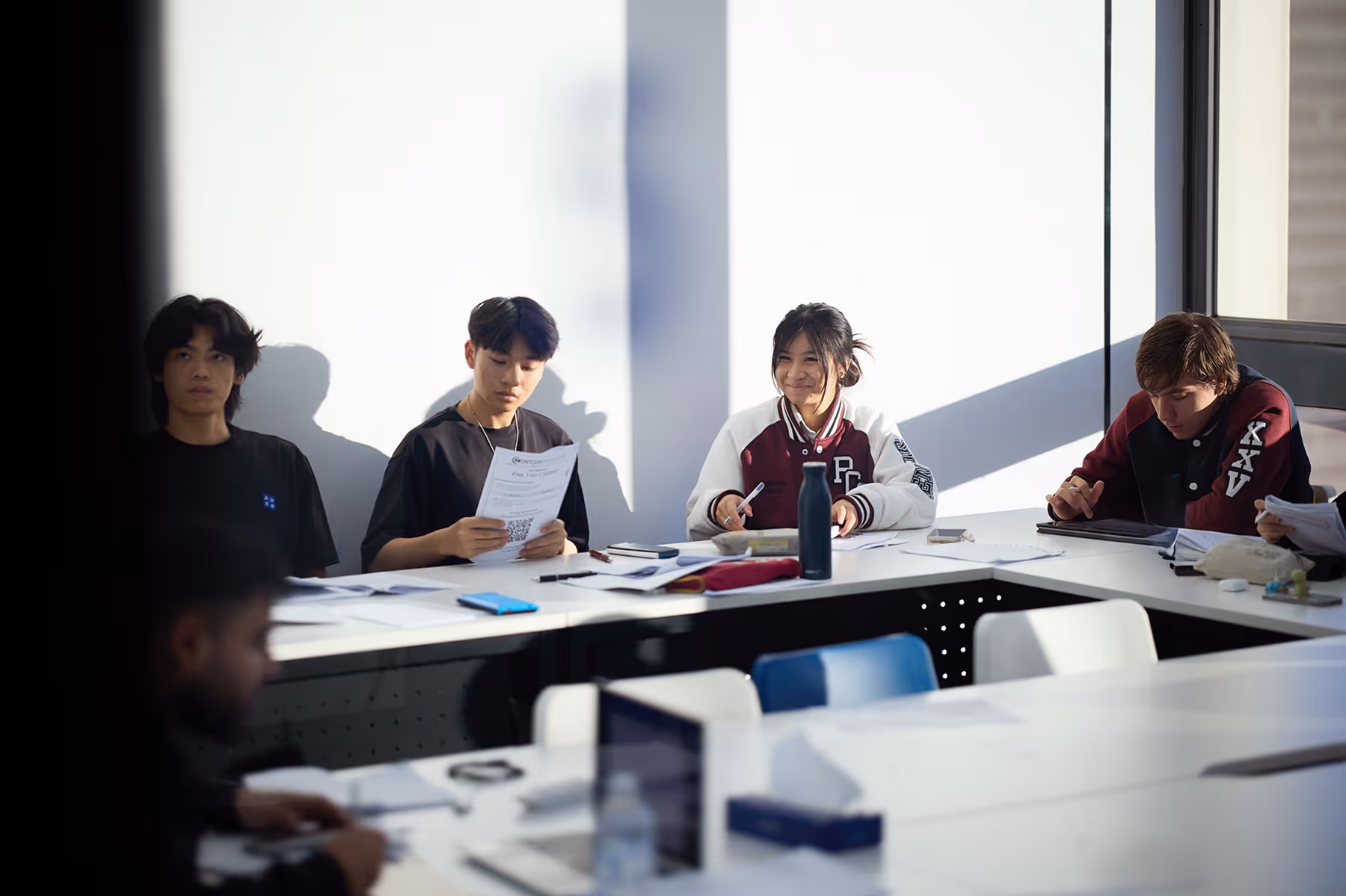 Group of young students sitting at a table with papers and laptops, studying in a sunlit classroom.