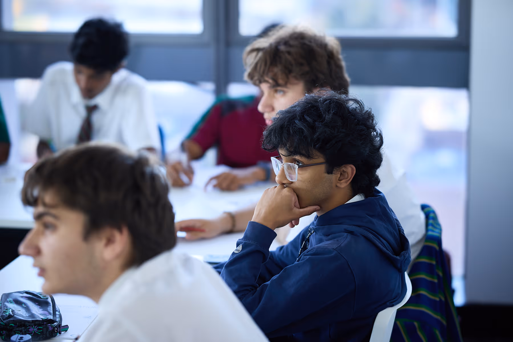 Students sitting at desks in a classroom, one wearing glasses and a blue hoodie appears thoughtful.