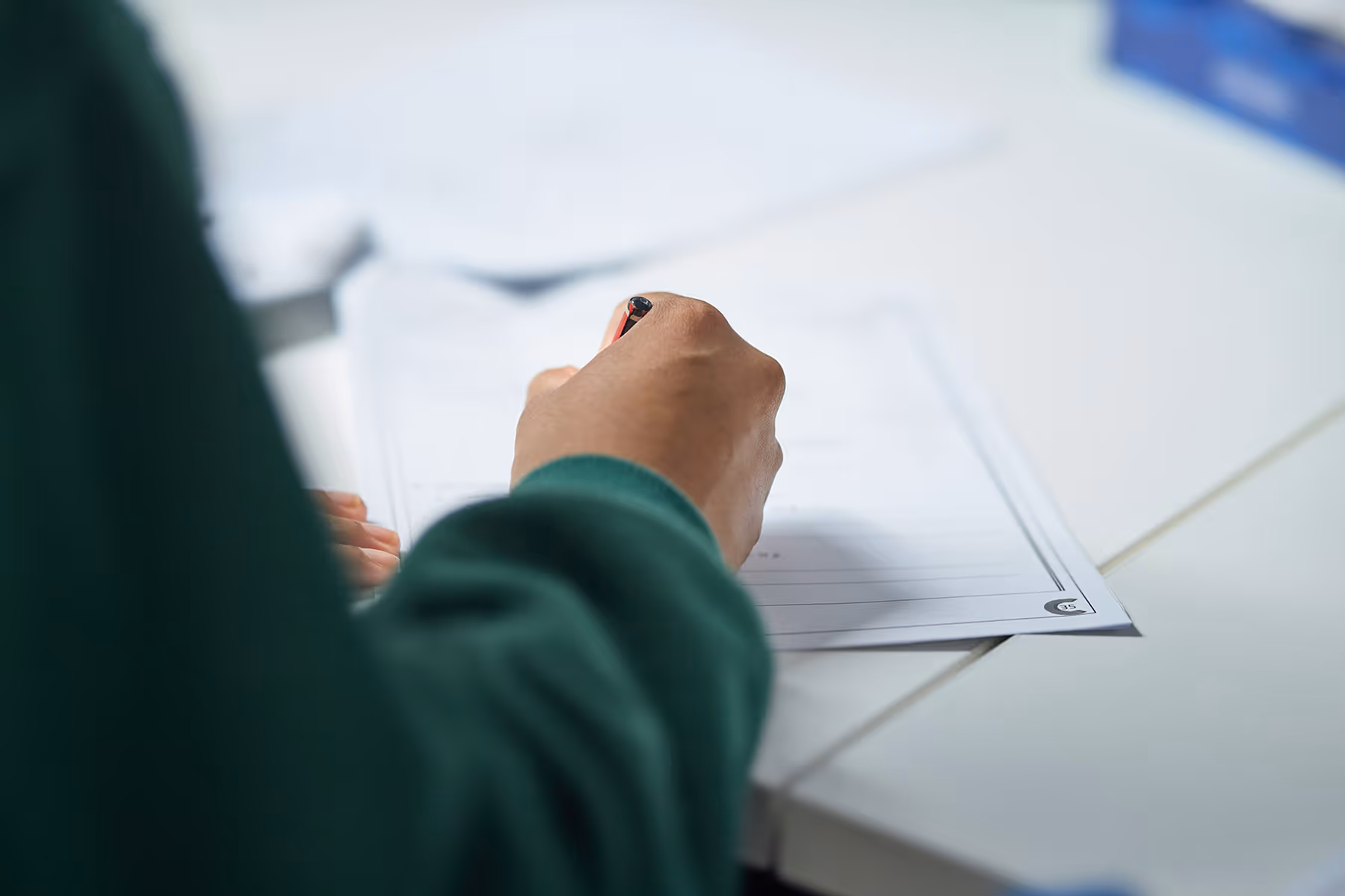 Person wearing a green sleeve writing on a sheet of paper with a pen on a white desk.