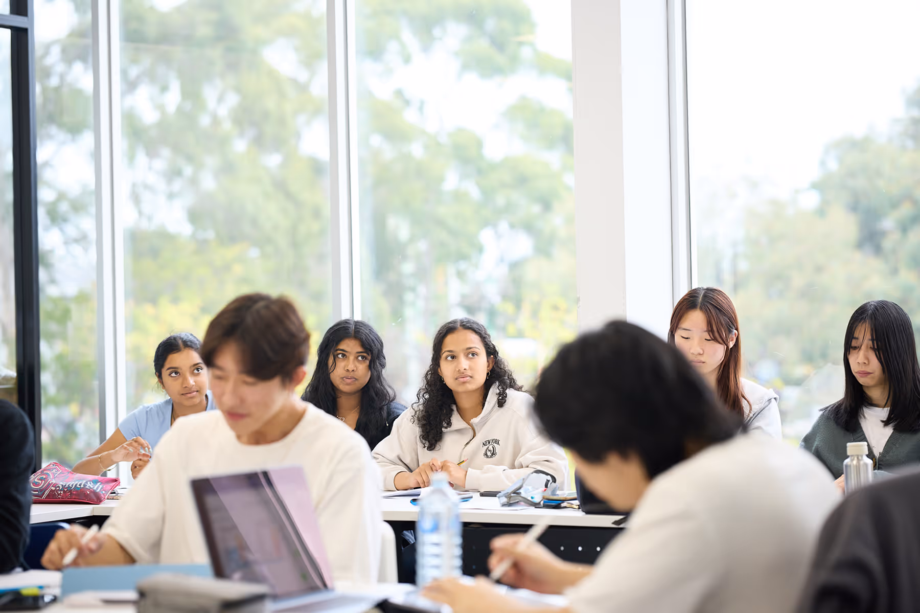 Students in a bright classroom study and take notes, with large windows showing green trees outside.