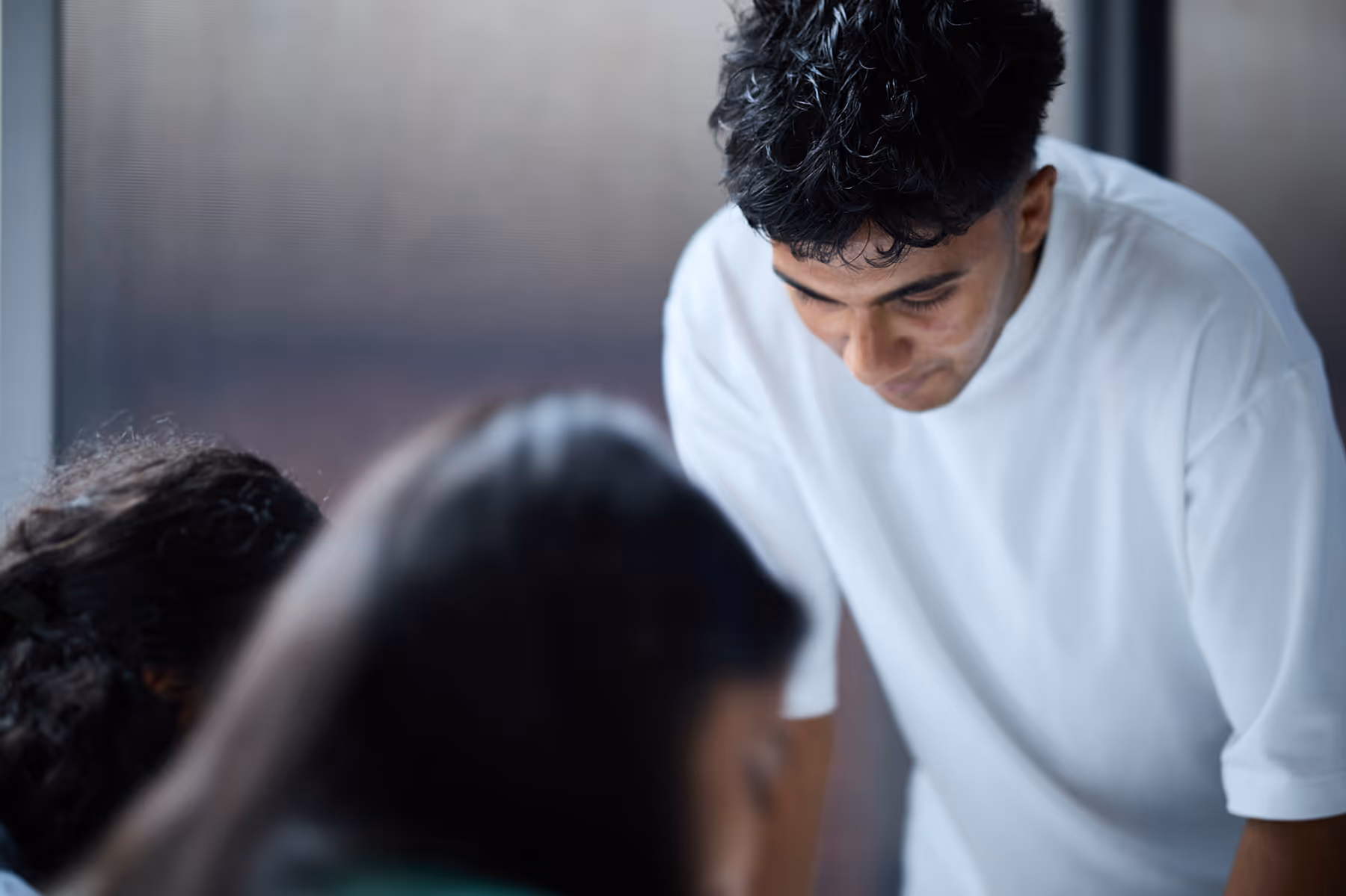 Young man in a white shirt leaning forward and looking down with two blurred people in the foreground.