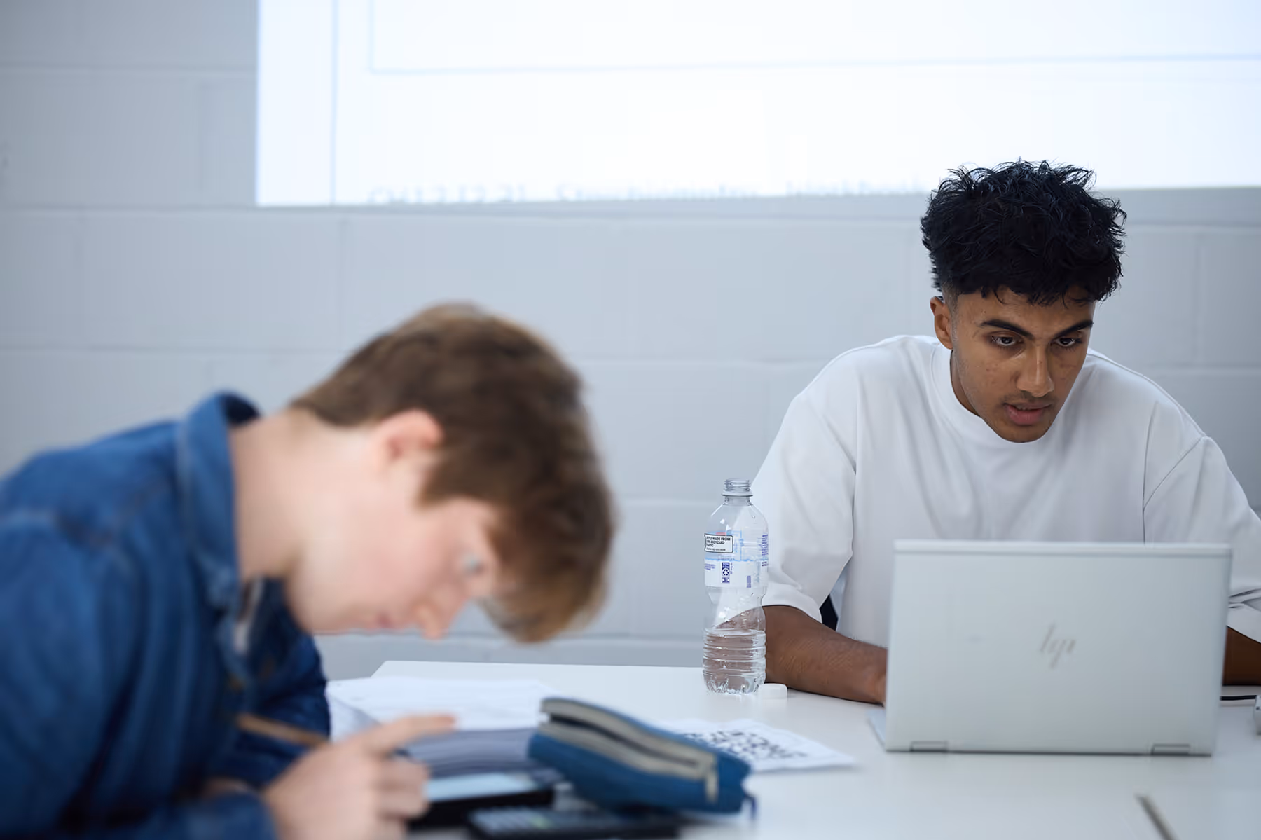 Two students studying at a white table, one focused on writing and the other looking at a laptop with a water bottle nearby.