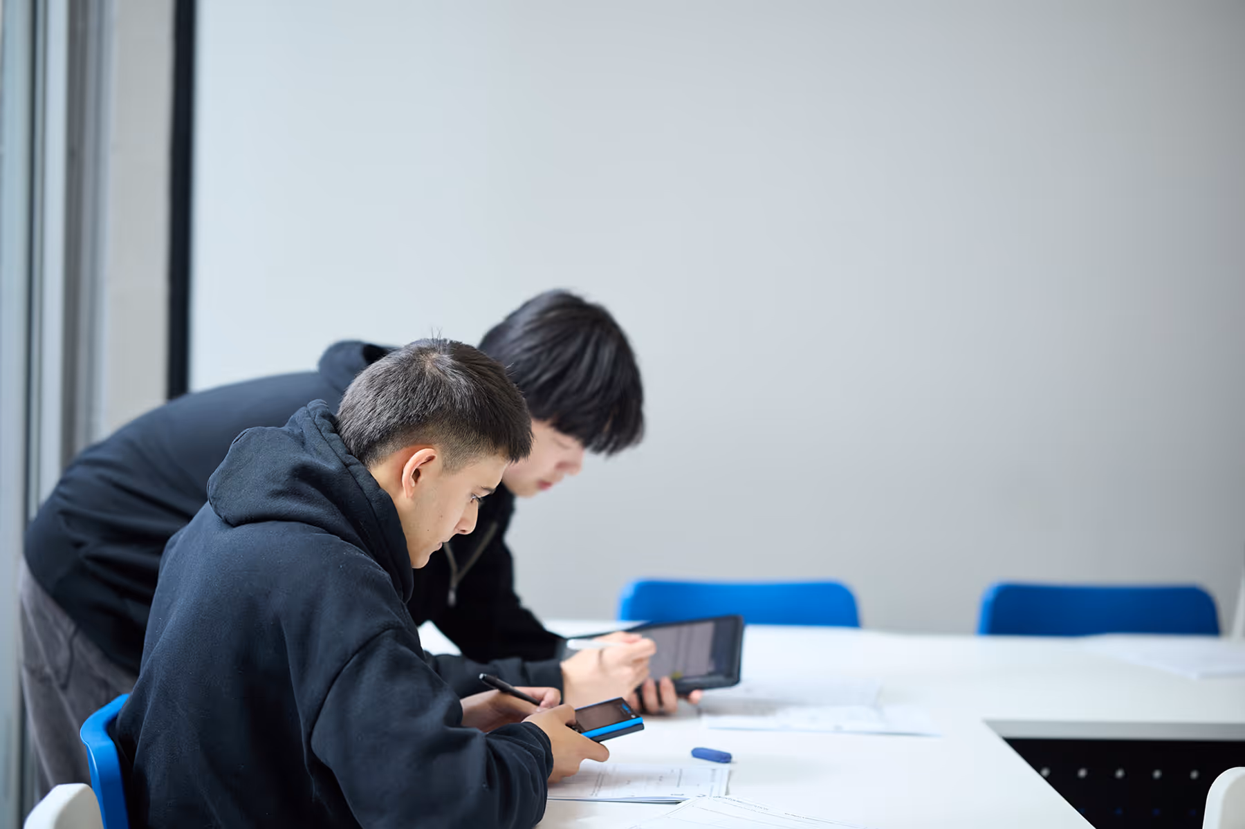 Three young men in black hoodies focused on studying with a smartphone and tablet at a white table in a classroom.