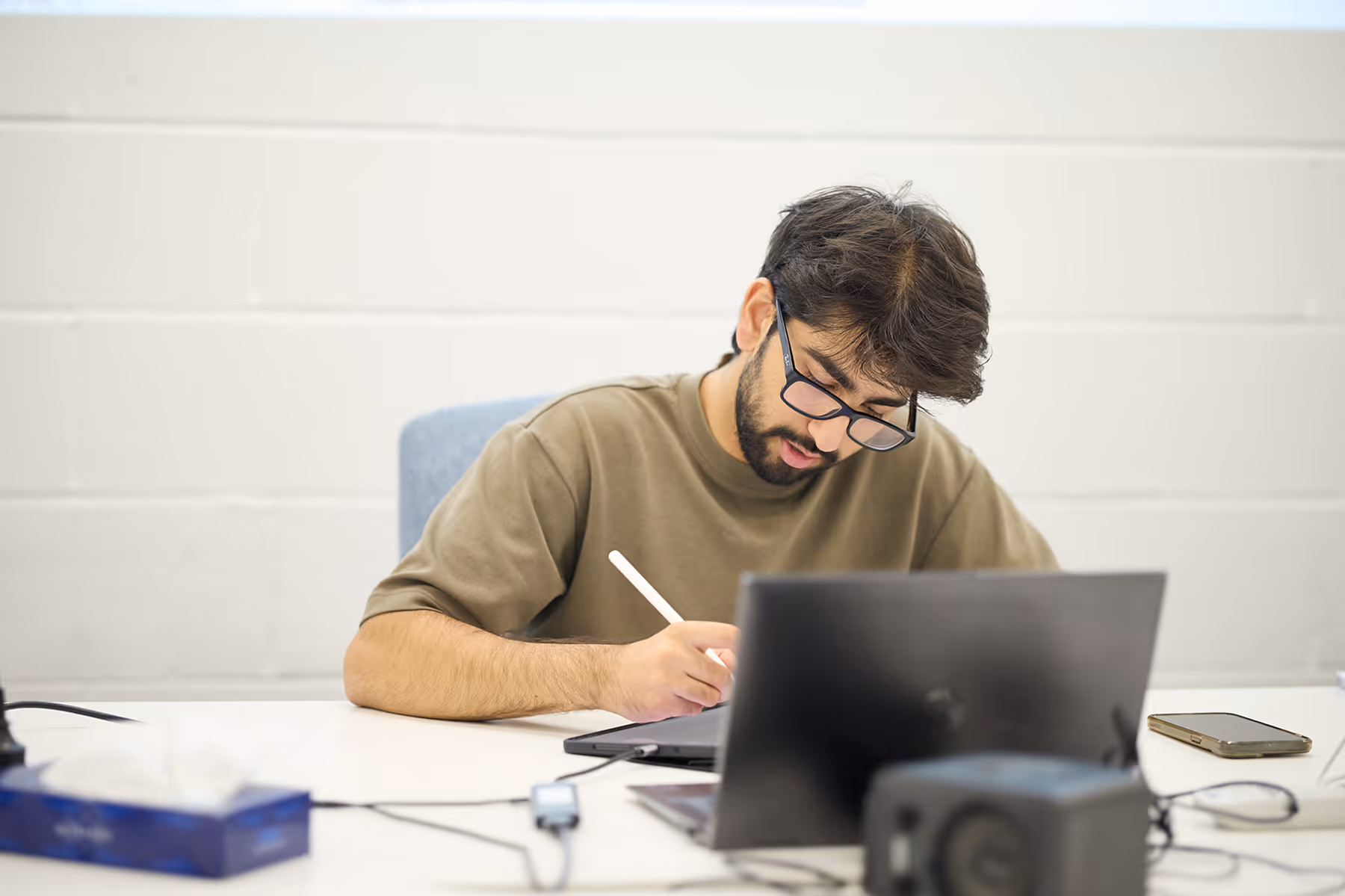 Man with glasses writing on a tablet while sitting at a desk with a laptop and smartphone.