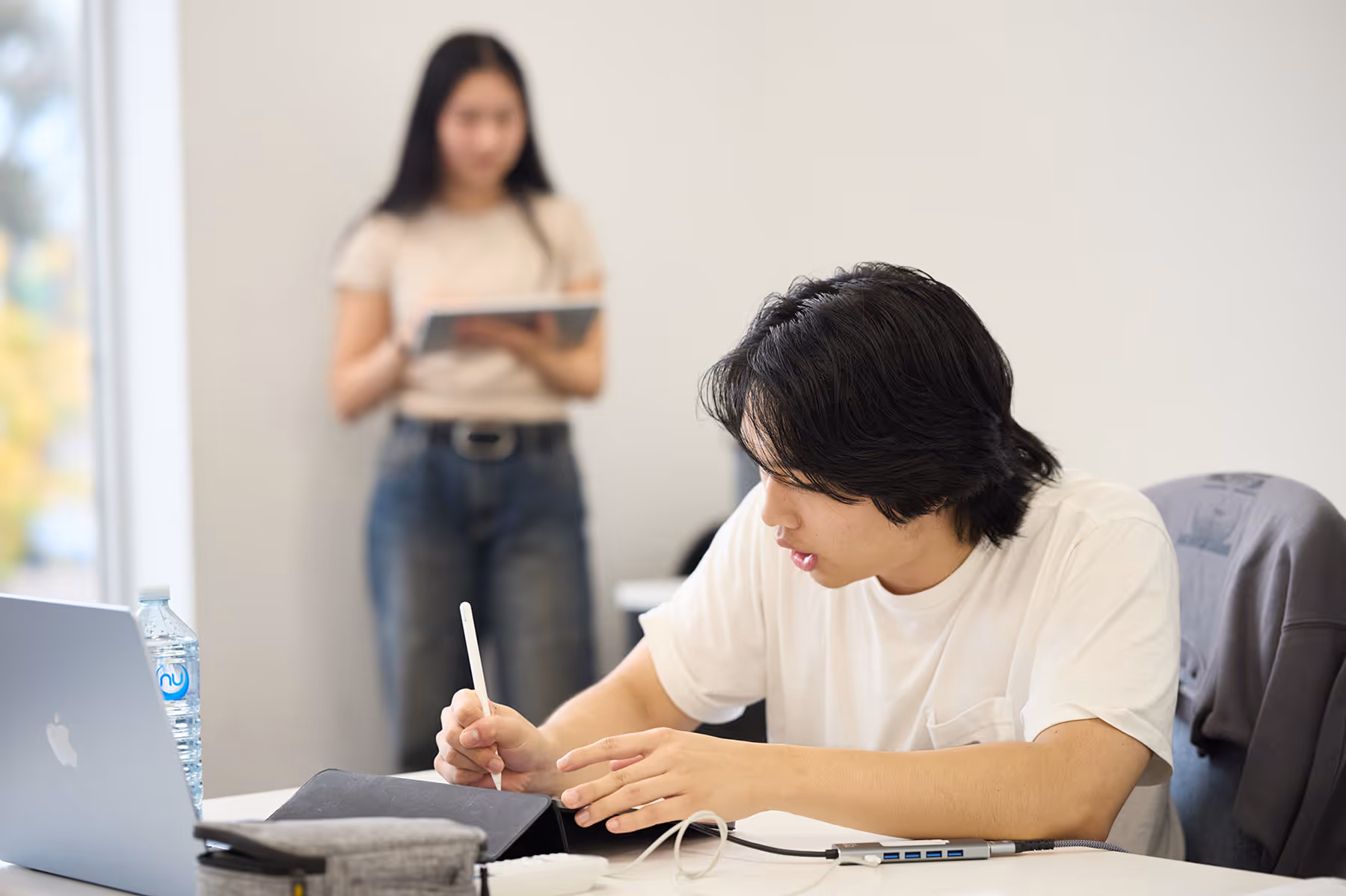 Young man in a white shirt using a digital pen on a tablet at a desk, with a laptop and water bottle nearby, and a woman standing blurred in the background holding a tablet.