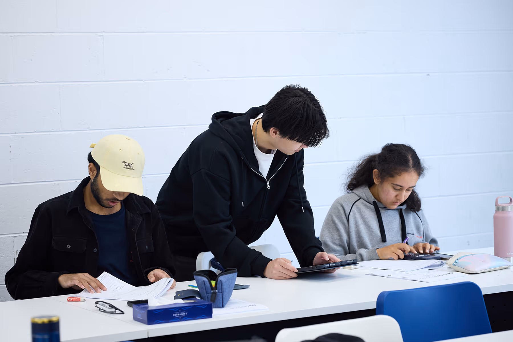 Three students studying at a white table with papers, a tablet, and a calculator in a classroom with a white brick wall.
