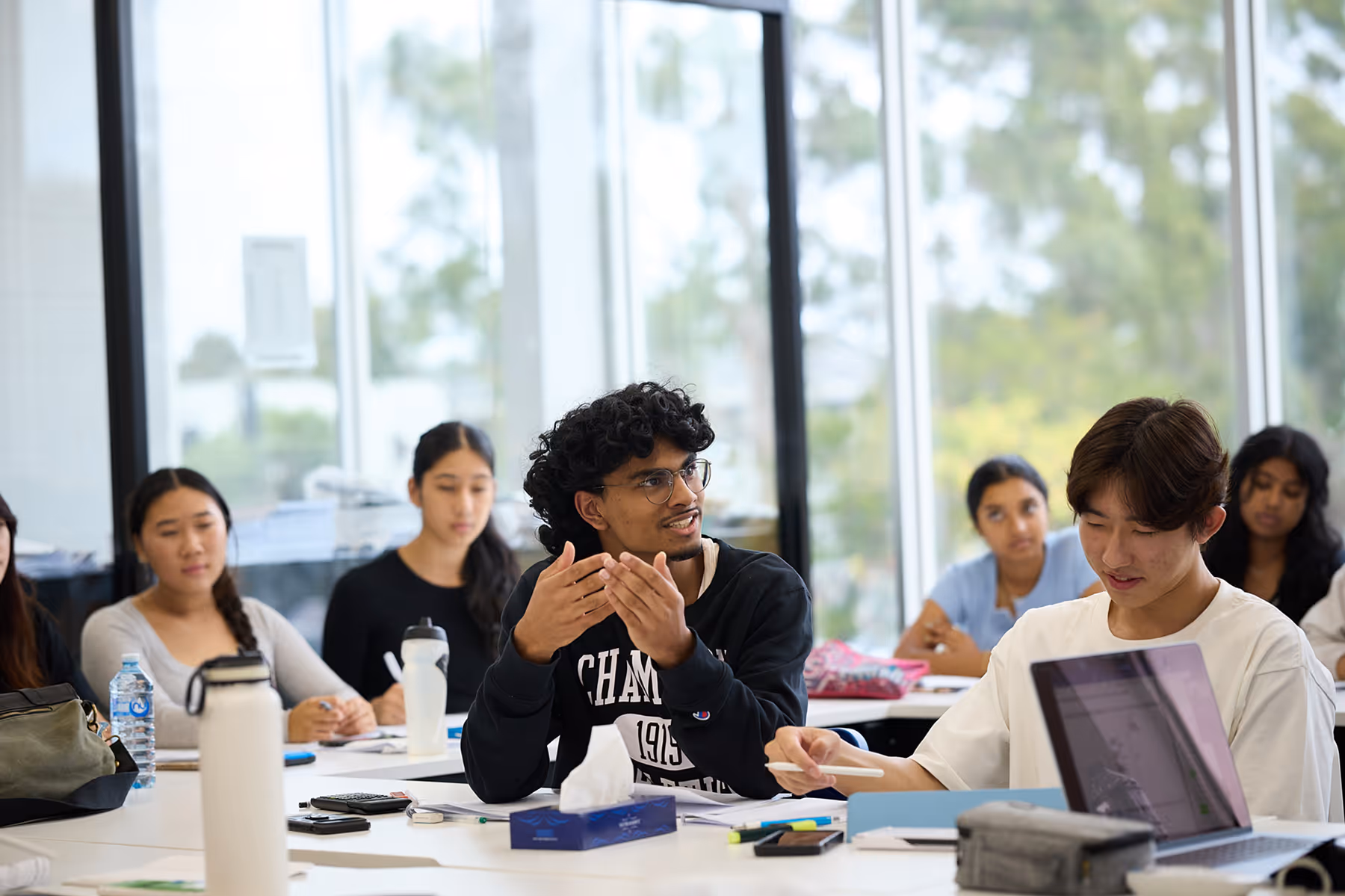 Students engaged in a classroom discussion, with one student speaking and others listening and working on laptops.