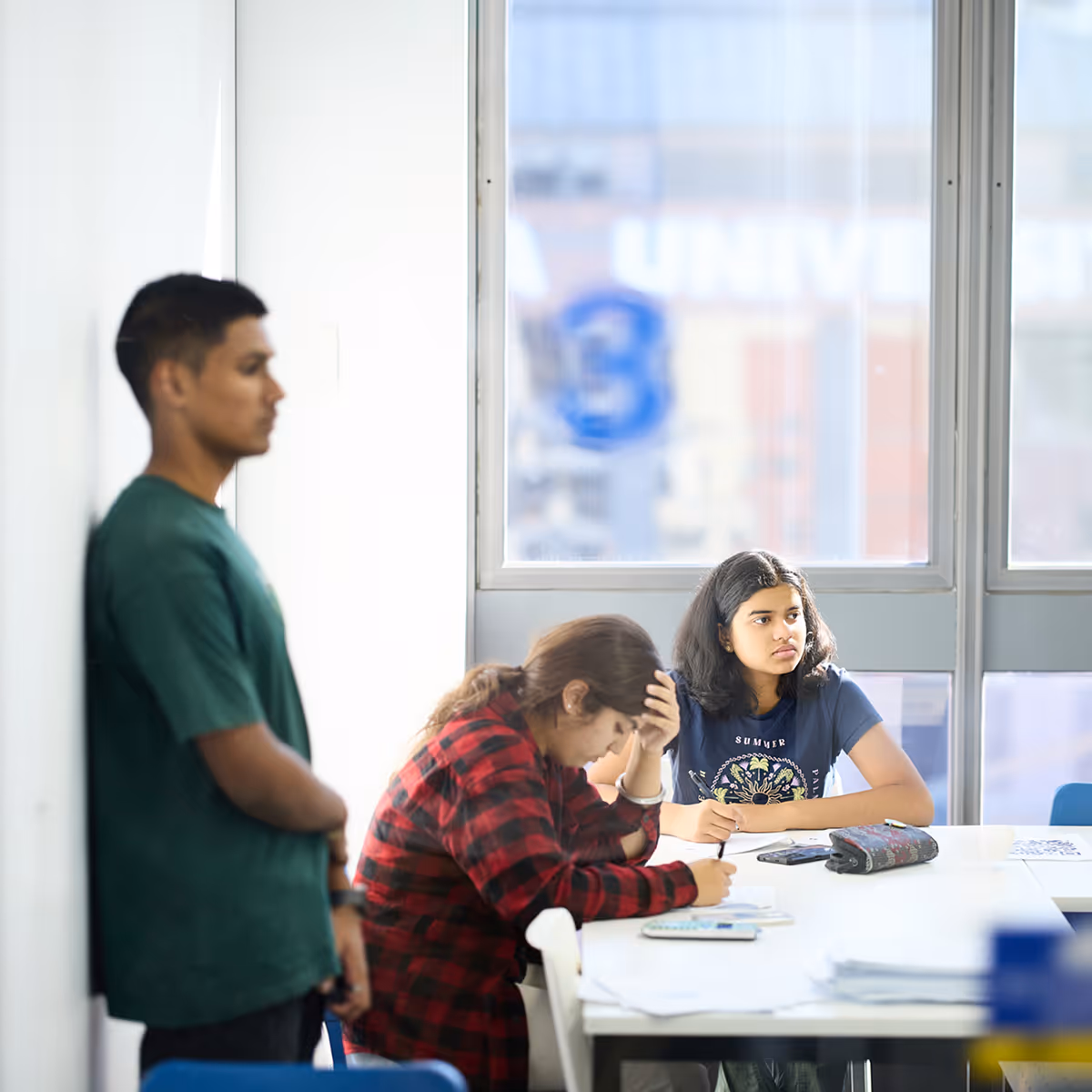 Two young women sitting at a table writing, while a young man in a green shirt stands nearby leaning against a wall.