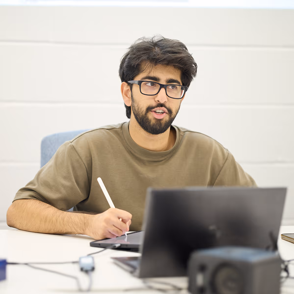 Young man with glasses and beard using a stylus on a tablet while sitting at a desk with a laptop.