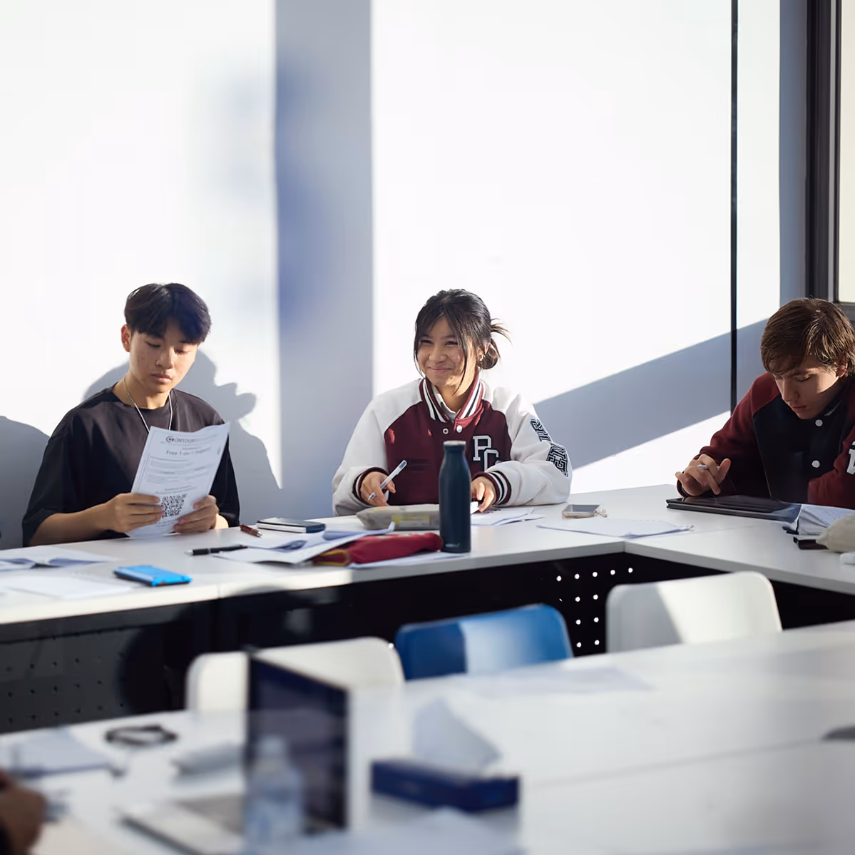 Three students sitting at a classroom table with papers and tablets, one smiling and holding a pen.
