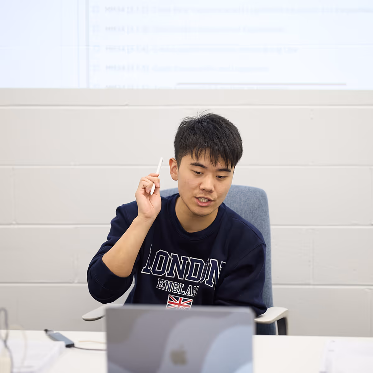 Young man in a London England sweatshirt holding a pen and speaking while sitting at a desk with a laptop.