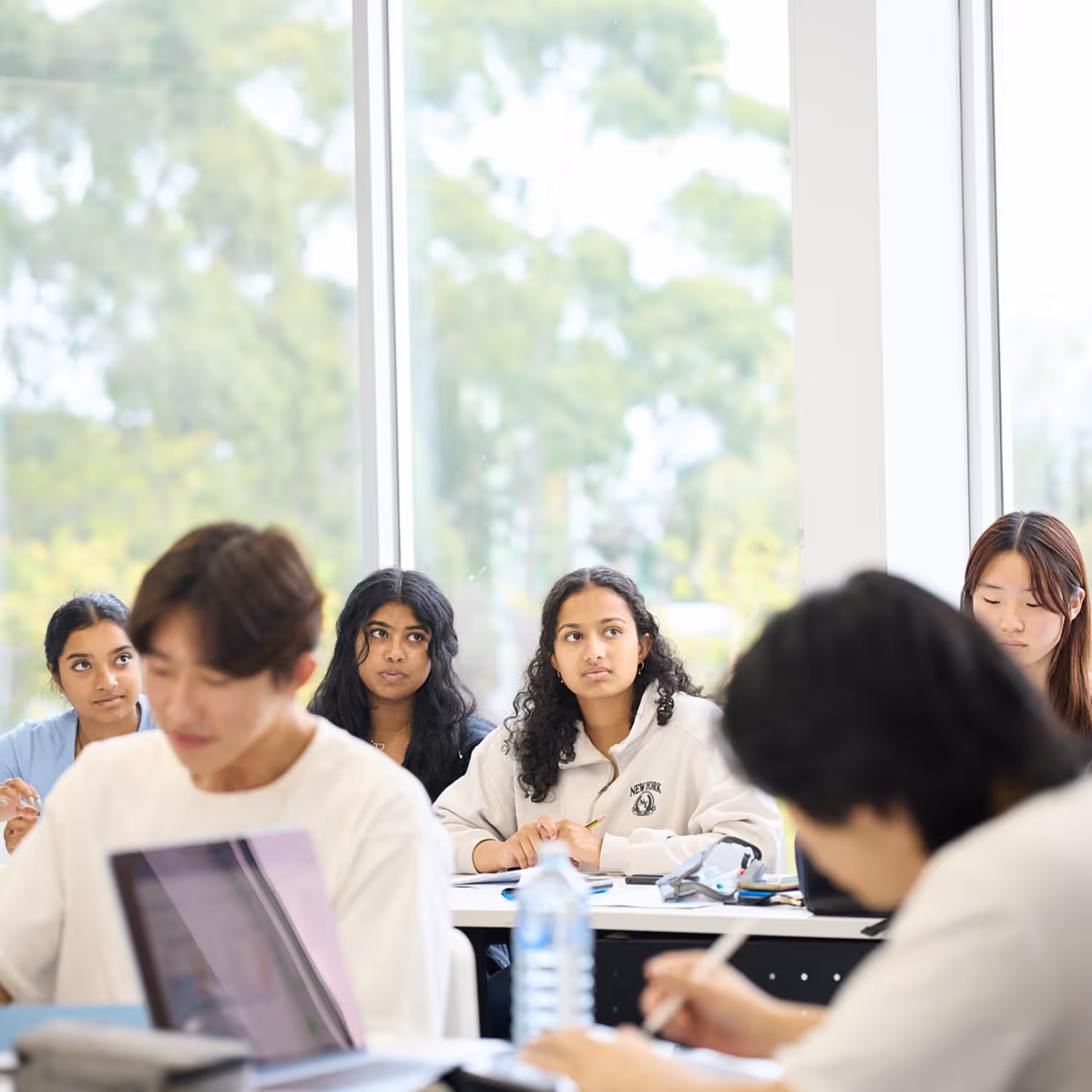 A group of diverse students sitting at desks in a classroom with large windows, some focused on writing and others looking attentive.