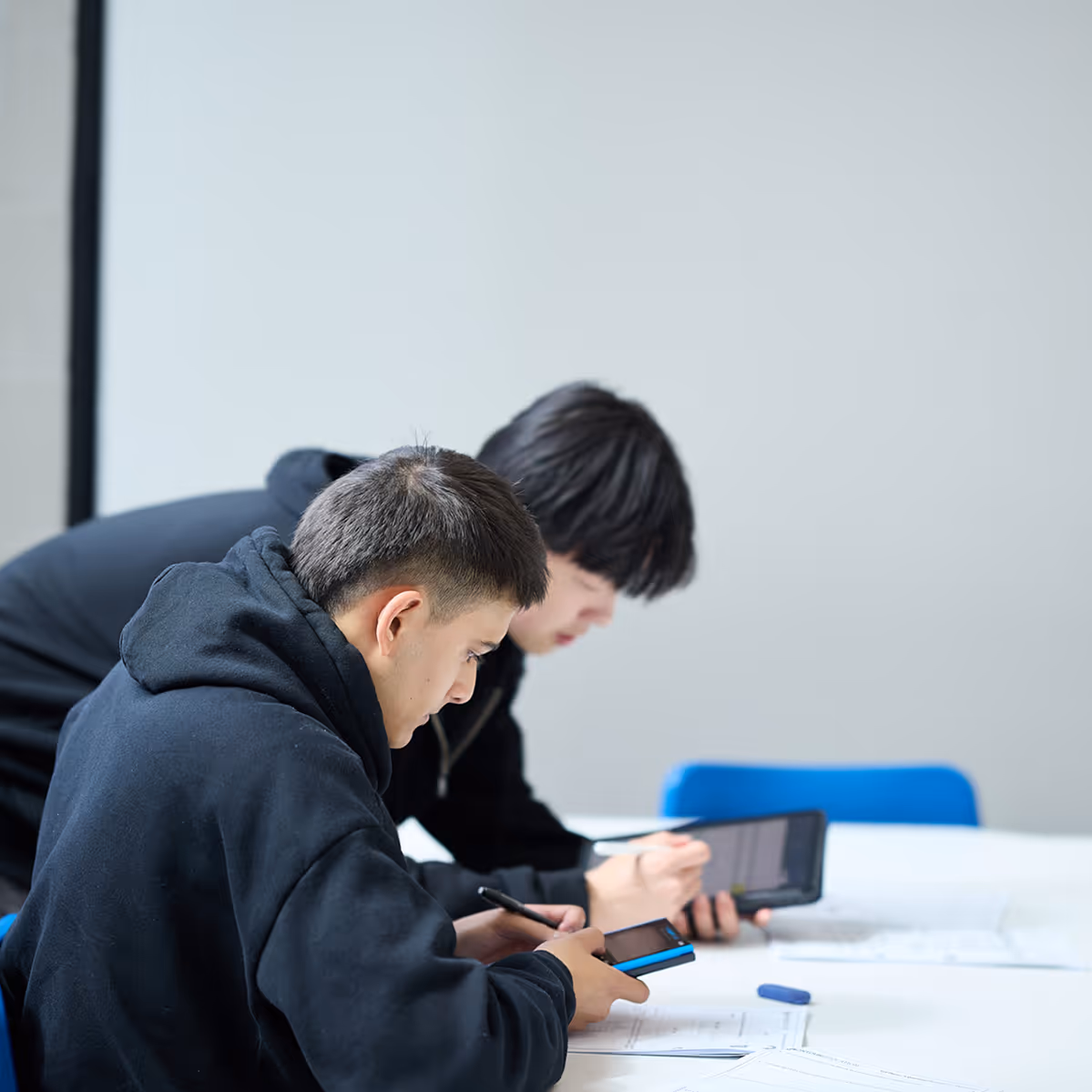 Two young men in black hoodies focused on using digital devices while sitting at a white table with papers.