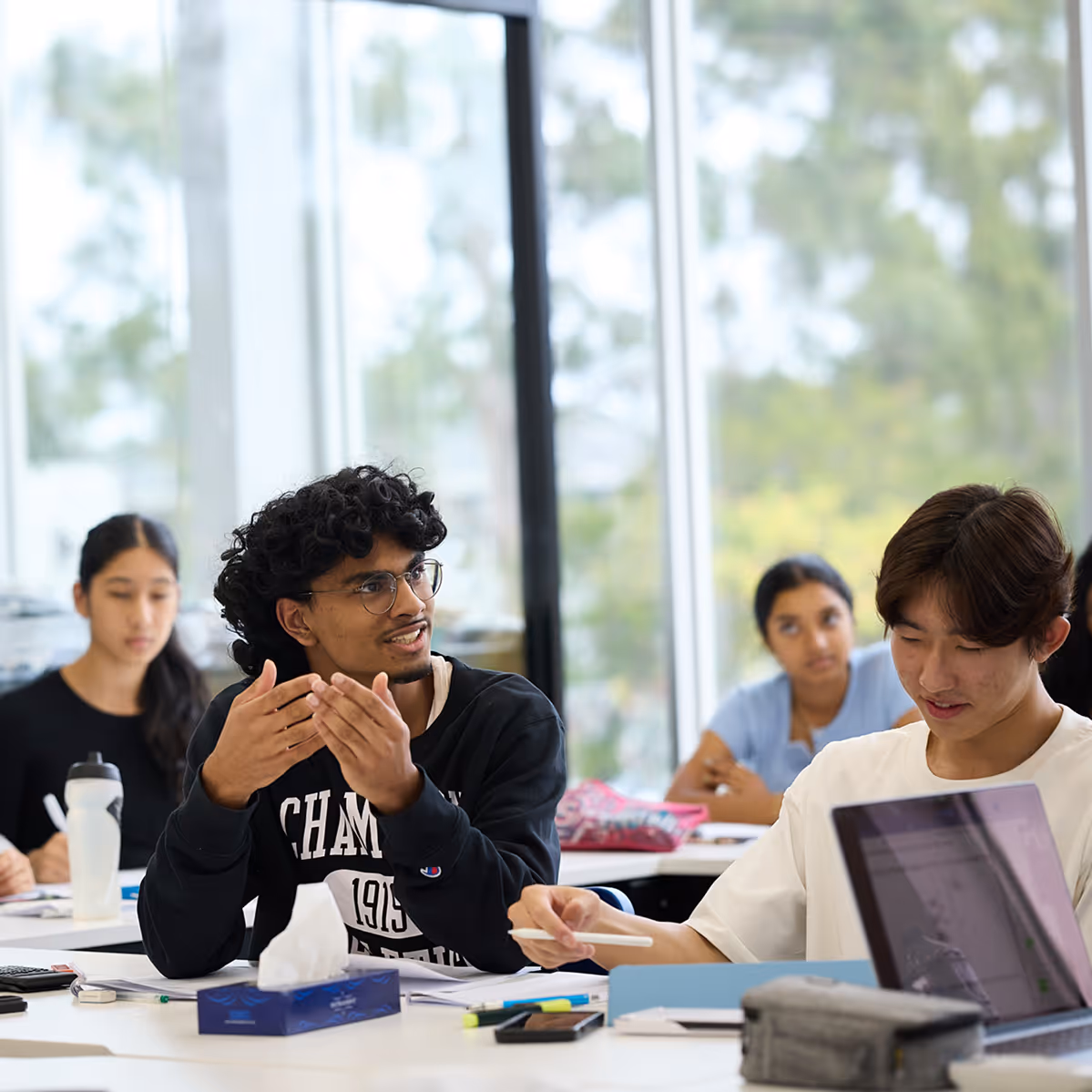 Students in a classroom engaged in discussion and working with study materials, including a laptop and stationery.