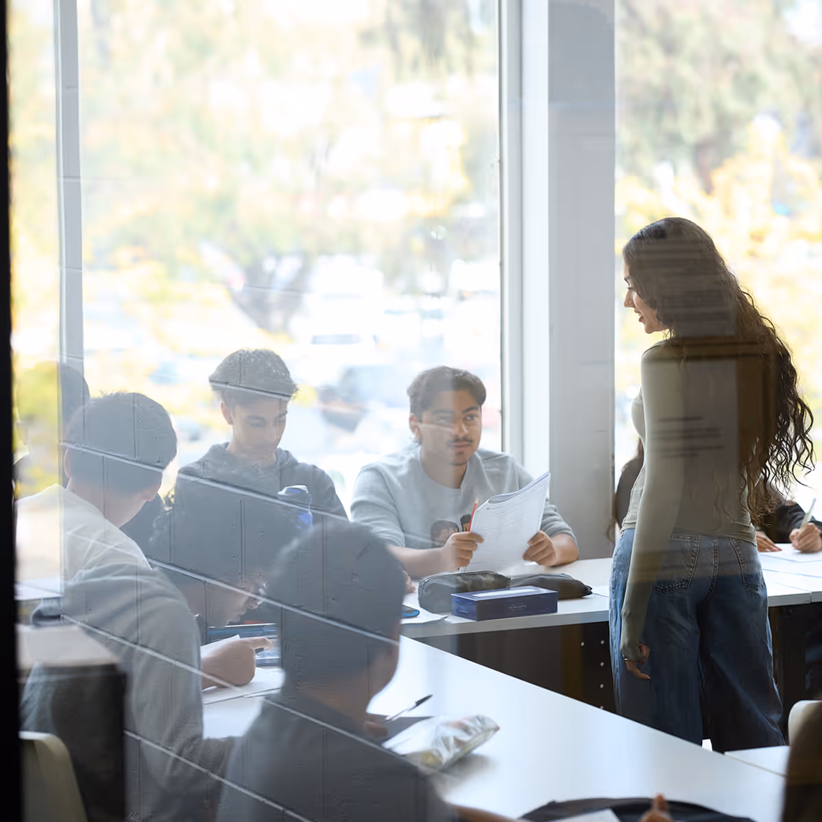 Group of students sitting and standing around a table in a classroom with large windows.