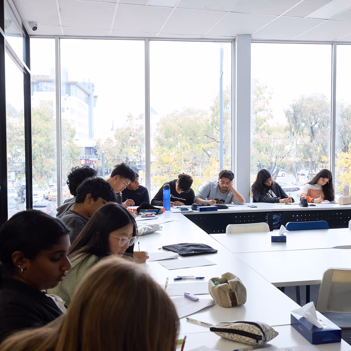 Group of students intensely writing or drawing on paper at white tables in a bright classroom with large windows.