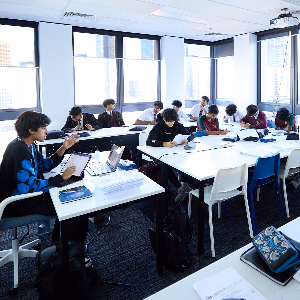Classroom with students seated at tables studying, some with laptops and papers, large windows with city view.