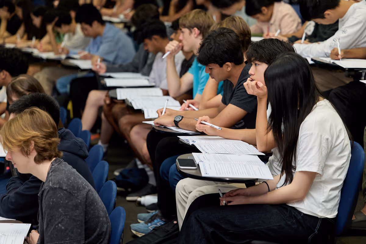 Students seated in a classroom writing on papers with pens on attached desks.
