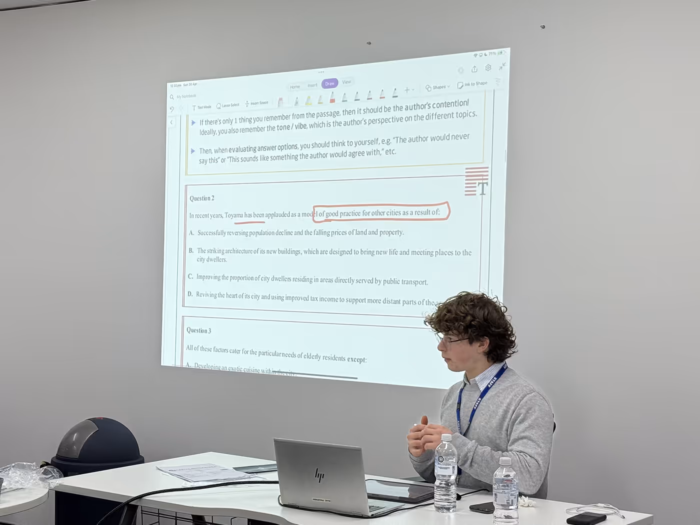 Person sitting at a table with a laptop and two water bottles, presenting a projected slide showing text about a question on good practice for cities with one phrase circled in red.